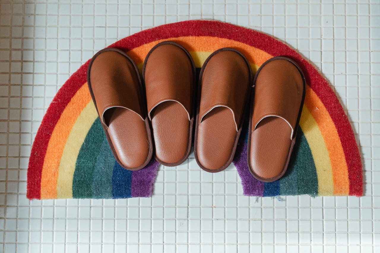 Top view of brown leather slippers on rainbow shaped colorful mat on white tiled bathroom floor